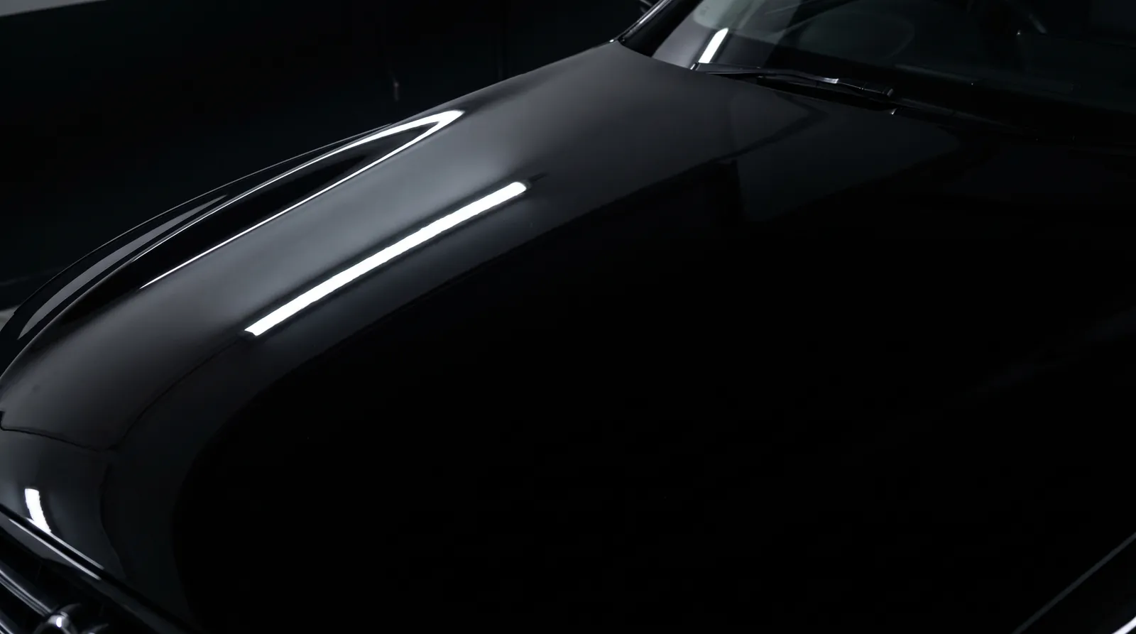 The same black bonnet after multi-stage correction, a defect-free mirror finish under the same lamp.