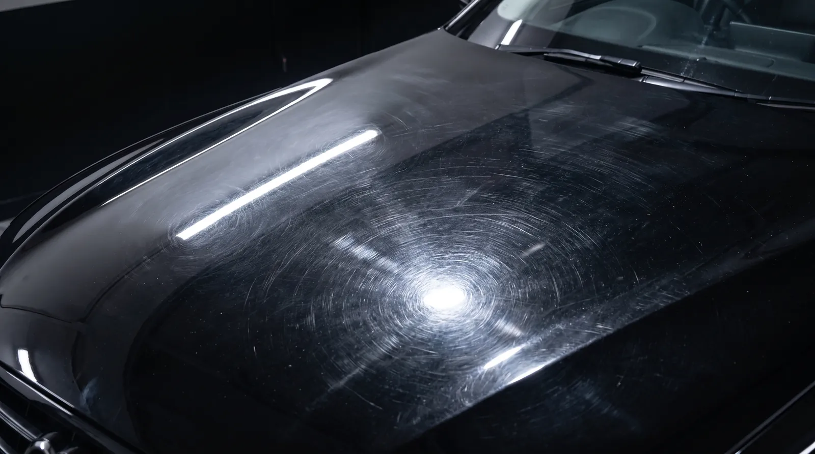 Black car bonnet covered in swirl marks and wash marring, lit by a raking inspection lamp.